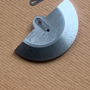 Pu&ograve; includere: Primo piano di componenti di orologi. Un componente metallico argentato a forma di semicerchio con un bordo nero e un piccolo pezzo rettangolare scuro sono mostrati su una superficie marrone. Il pezzo a semicerchio ha un foro.