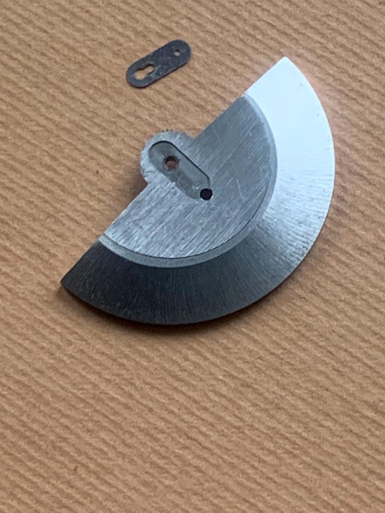 Pu&ograve; includere: Primo piano di componenti di orologi. Un componente metallico argentato a forma di semicerchio con un bordo nero e un piccolo pezzo rettangolare scuro sono mostrati su una superficie marrone. Il pezzo a semicerchio ha un foro.