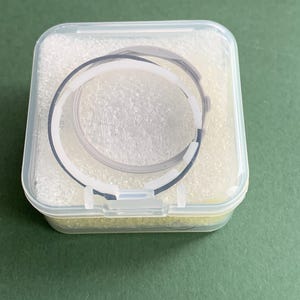 May include: A clear plastic case containing a black and white watch strap. The strap is circular and appears to be made of a flexible material. The case is filled with a white, foam-like material and sits on a green surface.