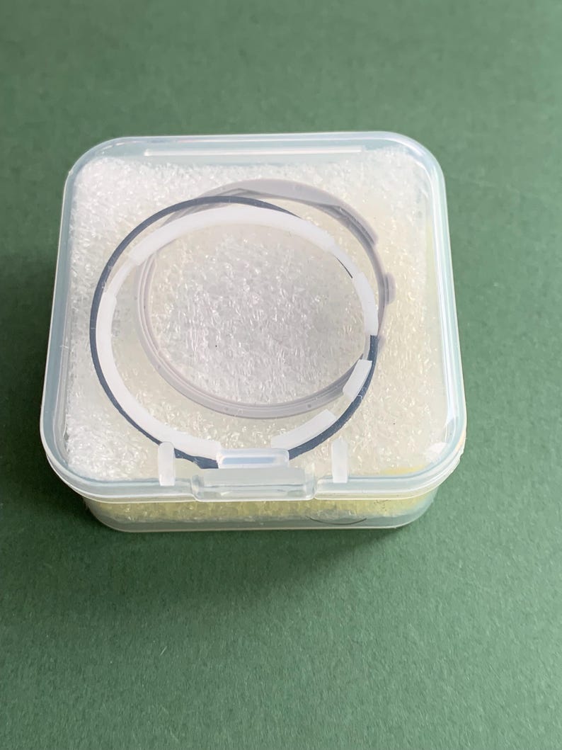 May include: A clear plastic case containing a black and white watch strap. The strap is circular and appears to be made of a flexible material. The case is filled with a white, foam-like material and sits on a green surface.