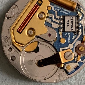 Może przedstawiać: Zbliżenie mechanizmu zegarka z misternymi detalami. Zawiera miedzianą cewkę, złote elementy i płytkę obwodu z napisem "CERTINA SWISS 729 V8". Mechanizm zegarka jest srebrny i niebieski.