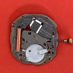 K&ouml;nnte beinhalten: Nahaufnahme eines Uhrwerks der Orient Watch Co. Ltd. Der silberfarbene Mechanismus hat eine Kupferspule, eine wei&szlig;e Batterie und eine goldfarbene Krone. Der Text "B6700 JAPAN" ist sichtbar.