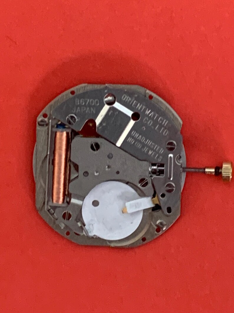 K&ouml;nnte beinhalten: Nahaufnahme eines Uhrwerks der Orient Watch Co. Ltd. Der silberfarbene Mechanismus hat eine Kupferspule, eine wei&szlig;e Batterie und eine goldfarbene Krone. Der Text "B6700 JAPAN" ist sichtbar.