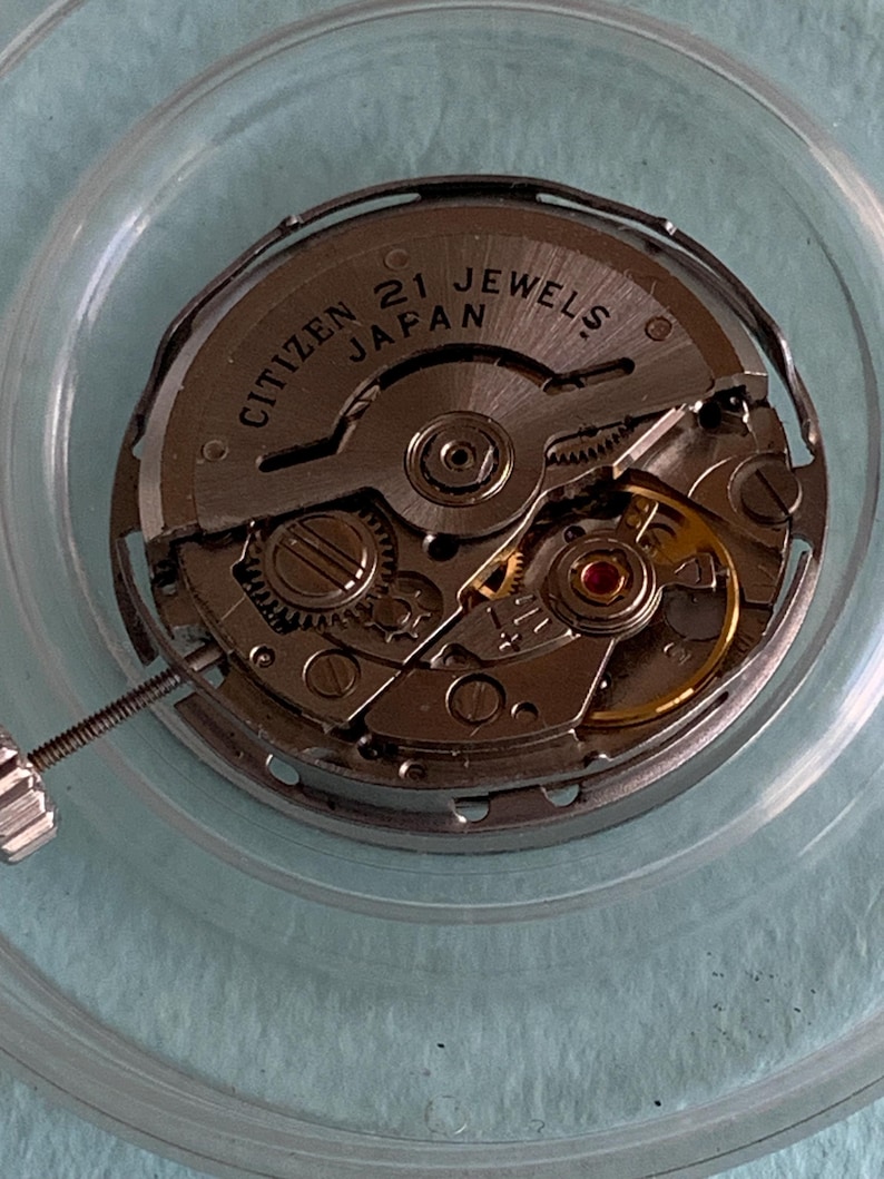 May include: A close-up of a Citizen watch movement with the text "CITIZEN 21 JEWELS JAPAN" visible. The watch mechanism is silver with gold-colored gears and a small screw. The movement is encased in a clear, circular container.