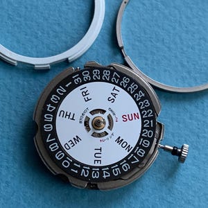 May include: A detailed view of a watch movement with a white dial and black numerals. The days of the week are visible, along with the date. Two silver rings are in the background, with a blue surface.