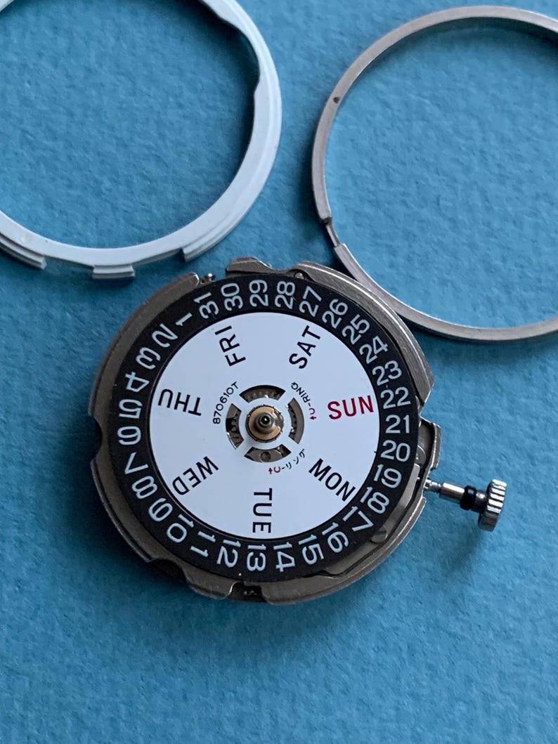 May include: A detailed view of a watch movement with a white dial and black numerals. The days of the week are visible, along with the date. Two silver rings are in the background, with a blue surface.