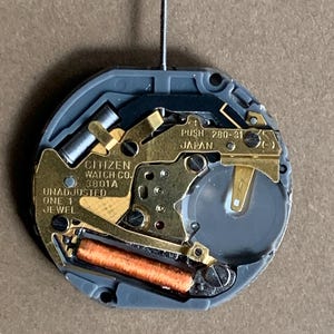 May include: A close-up of a Citizen watch movement. The movement features gold-colored metal components, a gray plastic casing, and a small battery. Text on the movement includes "CITIZEN WATCH CO.", "UNADJUSTED", and "JAPAN". A silver stem protrudes from the top.
