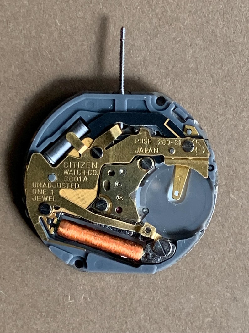 May include: A close-up of a Citizen watch movement. The movement features gold-colored metal components, a gray plastic casing, and a small battery. Text on the movement includes "CITIZEN WATCH CO.", "UNADJUSTED", and "JAPAN". A silver stem protrudes from the top.