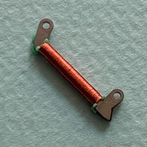 May include: A small, angled electronic component featuring a coil of red wire and two brown metal tabs with holes. Green insulation is visible at the coil's ends. The component is set against a light blue background.