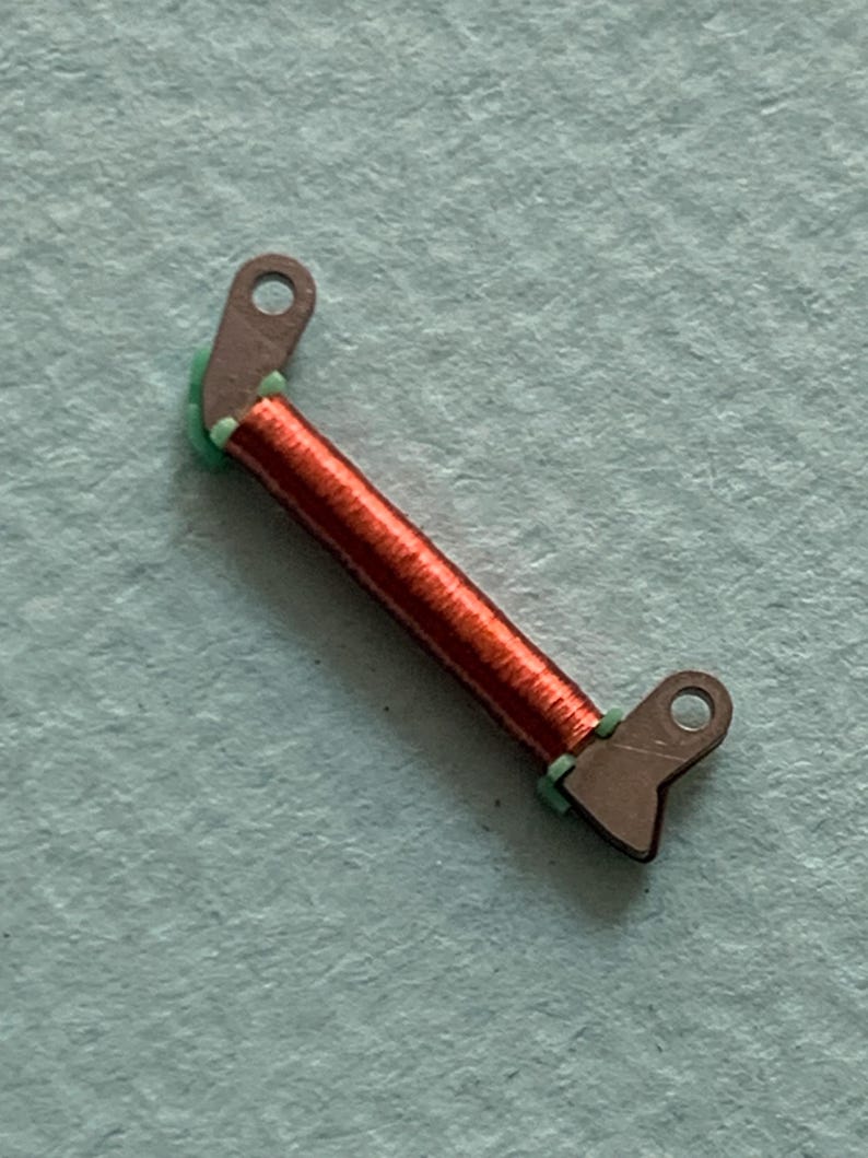 May include: A small, angled electronic component featuring a coil of red wire and two brown metal tabs with holes. Green insulation is visible at the coil's ends. The component is set against a light blue background.