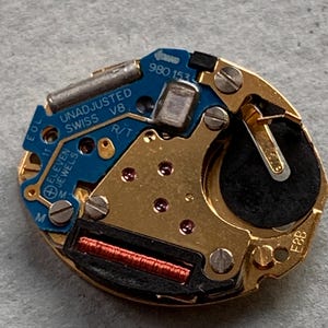 May include: A detailed view of a watch movement, featuring gold-toned metal parts and a blue circuit board. The board is marked with "UNADJUSTED SWISS V8" and "ELEVEN JEWELS". A black battery is also present.