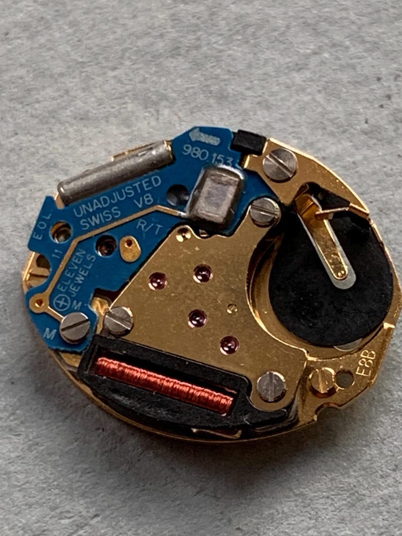May include: A detailed view of a watch movement, featuring gold-toned metal parts and a blue circuit board. The board is marked with "UNADJUSTED SWISS V8" and "ELEVEN JEWELS". A black battery is also present.