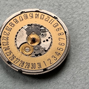 May include: A close-up of a watch movement with a gold-colored date wheel and intricate gears. The date wheel is marked with numbers 1 through 31. The watch mechanism is set in a silver-colored metal casing.