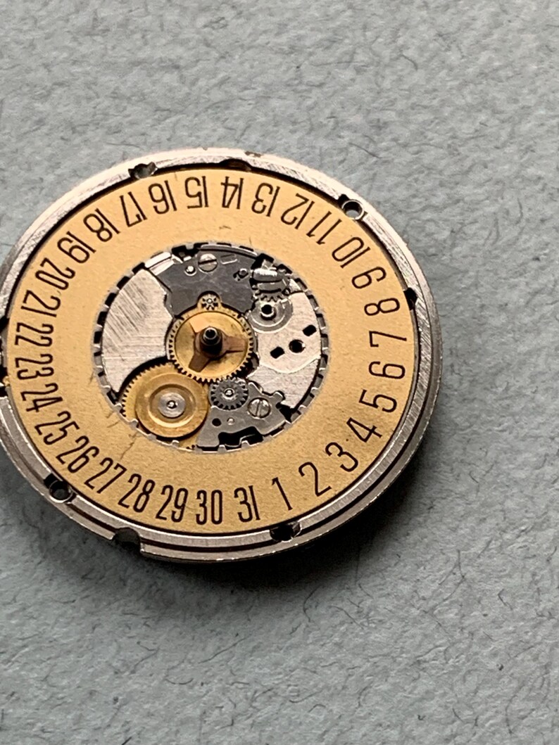 May include: A close-up of a watch movement with a gold-colored date wheel and intricate gears. The date wheel is marked with numbers 1 through 31. The watch mechanism is set in a silver-colored metal casing.