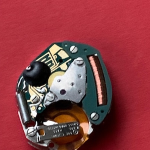 May include: A close-up of a watch movement on a red background. The movement features a green circuit board, silver metal components, and a gold-colored coil. The text "SWISS ADJUSTED" is visible on a small metal plate.
