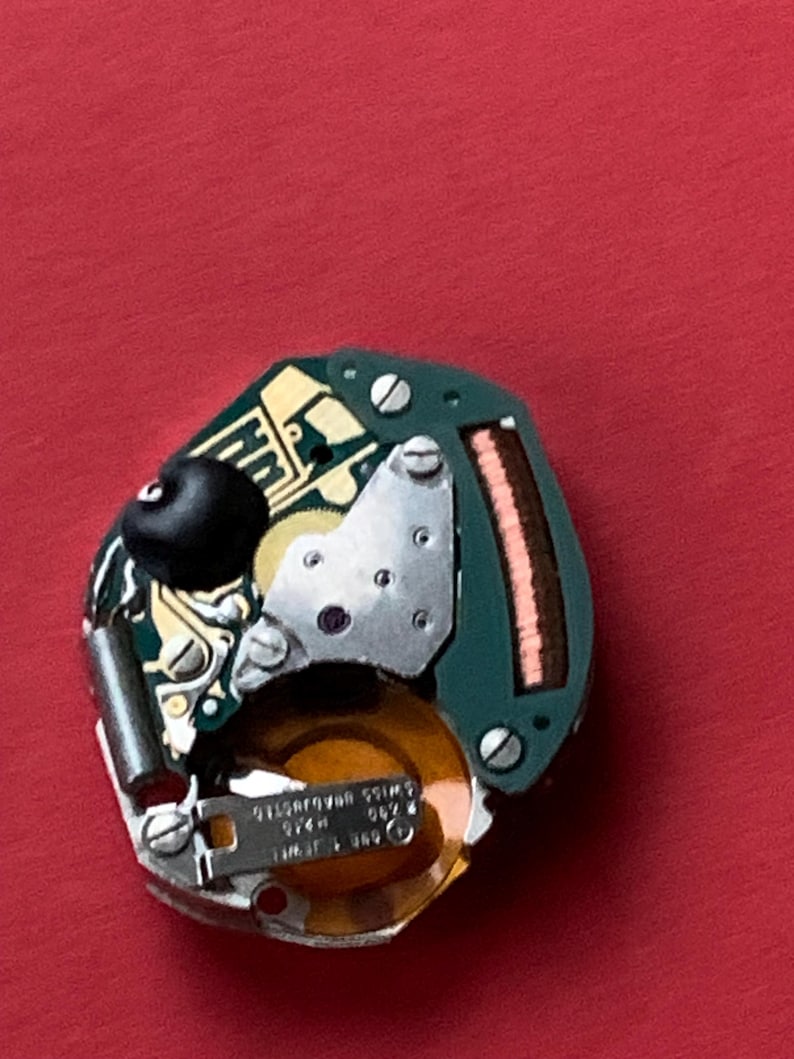 May include: A close-up of a watch movement on a red background. The movement features a green circuit board, silver metal components, and a gold-colored coil. The text "SWISS ADJUSTED" is visible on a small metal plate.