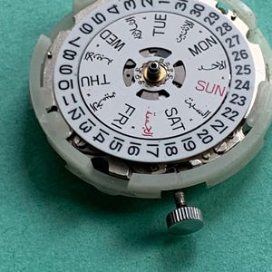 May include: Close-up of a watch movement with a white dial featuring Arabic and English day and date indicators. The dial has a central gold-colored component and a silver crown. The background is a solid teal color.