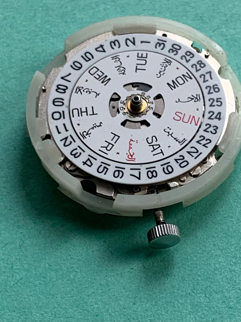 May include: Close-up of a watch movement with a white dial featuring Arabic and English day and date indicators. The dial has a central gold-colored component and a silver crown. The background is a solid teal color.