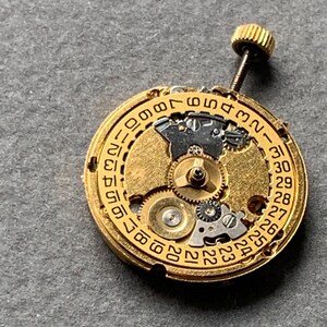 May include: Close-up of a gold-toned watch movement with intricate gears and a date dial. The dial displays numbers from 1 to 31. A small, threaded crown extends from the top. The background is a neutral gray.
