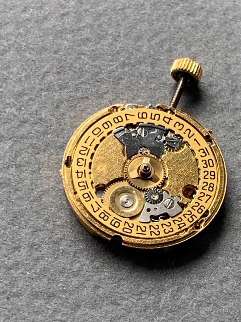 May include: Close-up of a gold-toned watch movement with intricate gears and a date dial. The dial displays numbers from 1 to 31. A small, threaded crown extends from the top. The background is a neutral gray.