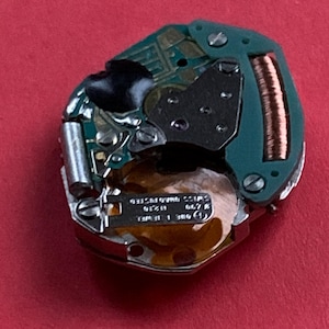 May include: A close-up of a watch movement with a green circuit board, metal components, and a copper coil. The watch mechanism has small screws and a gold-colored base. The background is a solid red.