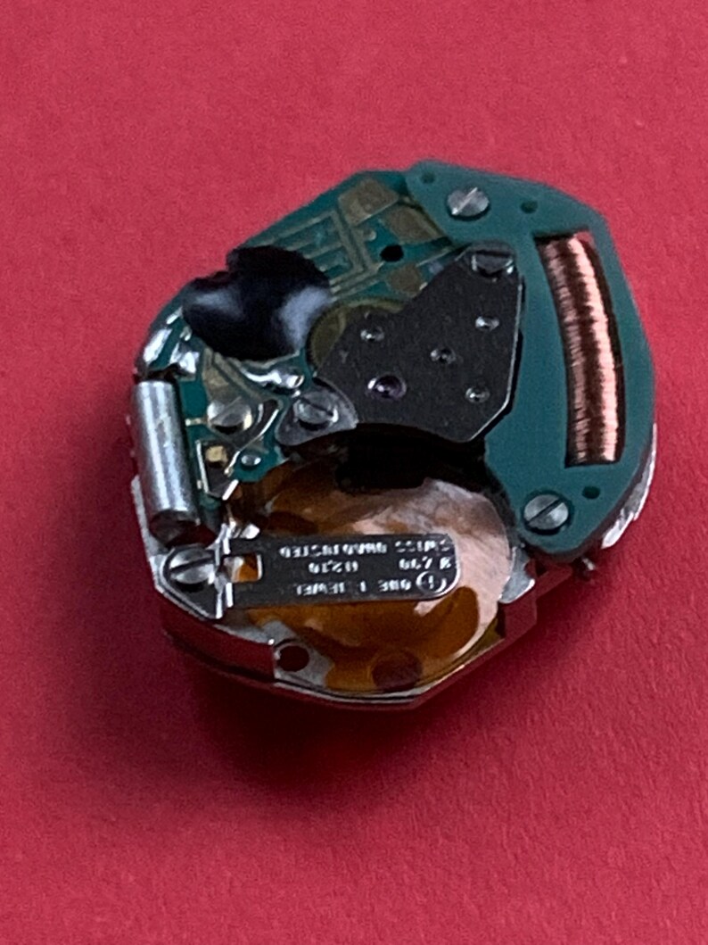 May include: A close-up of a watch movement with a green circuit board, metal components, and a copper coil. The watch mechanism has small screws and a gold-colored base. The background is a solid red.