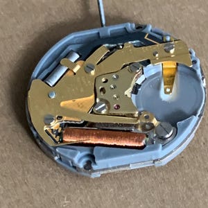 May include: Close-up of a watch movement with gold-colored gears, a copper-colored coil, and a silver-colored battery. The movement is housed in a gray plastic casing and has a small, silver post extending upwards.