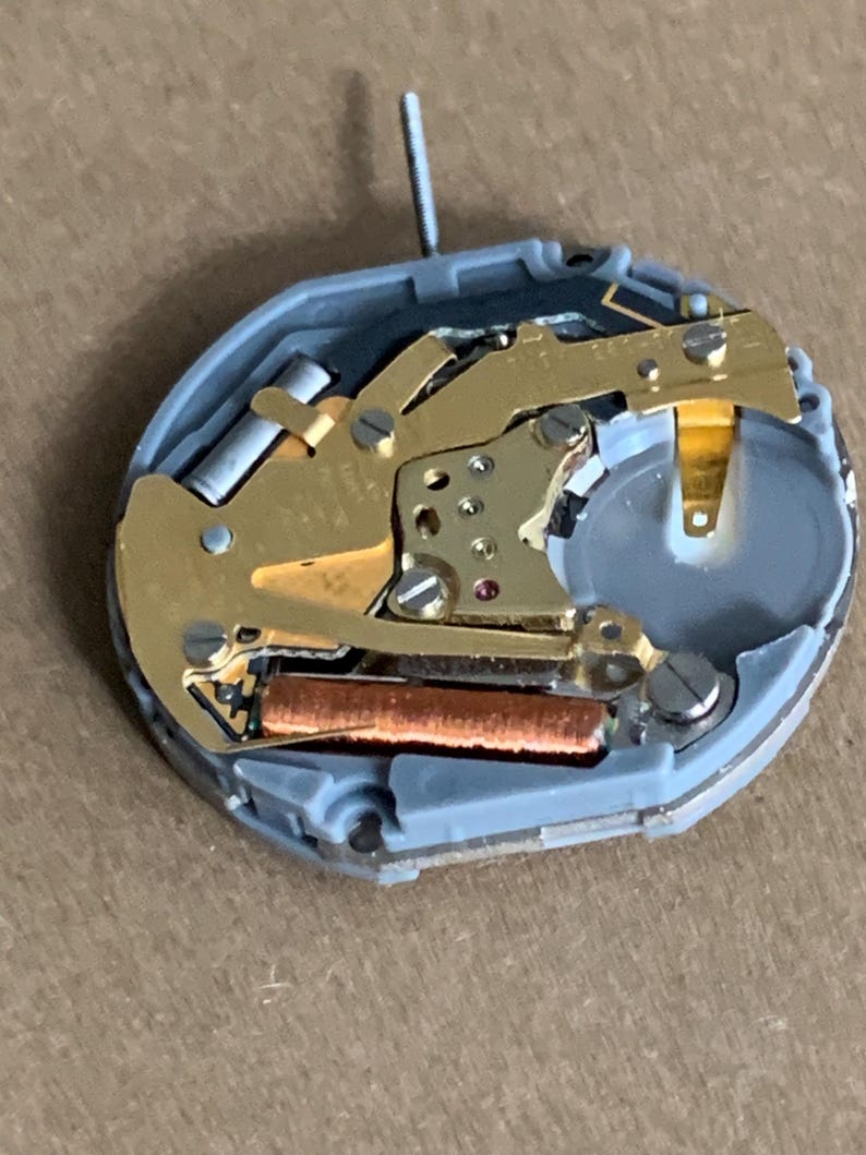 May include: Close-up of a watch movement with gold-colored gears, a copper-colored coil, and a silver-colored battery. The movement is housed in a gray plastic casing and has a small, silver post extending upwards.