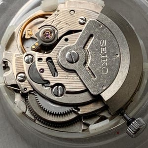 May include: Close-up of a Seiko watch movement, showcasing intricate gears, screws, and components. The silver-toned metal parts are arranged within a clear, circular casing. The word "SEIKO" is visible on a metal plate.