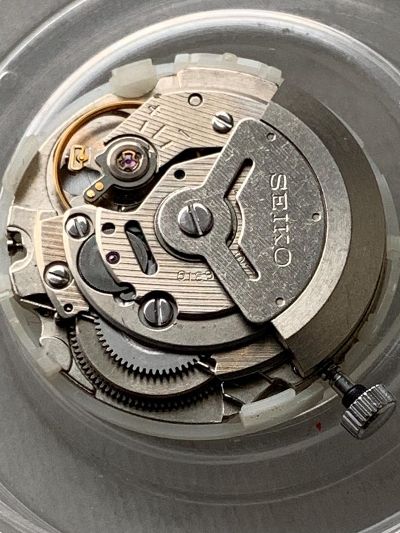 May include: Close-up of a Seiko watch movement, showcasing intricate gears, screws, and components. The silver-toned metal parts are arranged within a clear, circular casing. The word "SEIKO" is visible on a metal plate.