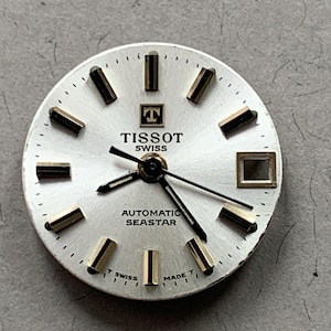 May include: A silver watch face with the brand name "TISSOT SWISS" and the words "AUTOMATIC SEASTAR" printed on it. The face has gold-coloured rectangular hour markers and black hands.