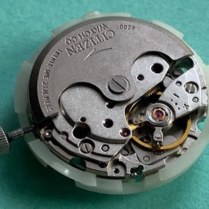 May include: Close-up of a Citizen watch movement with visible gears, jewels, and the text "Citizen Watch Co." The movement is silver-toned with a white plastic casing and a small screw. The watch movement is on a teal surface.