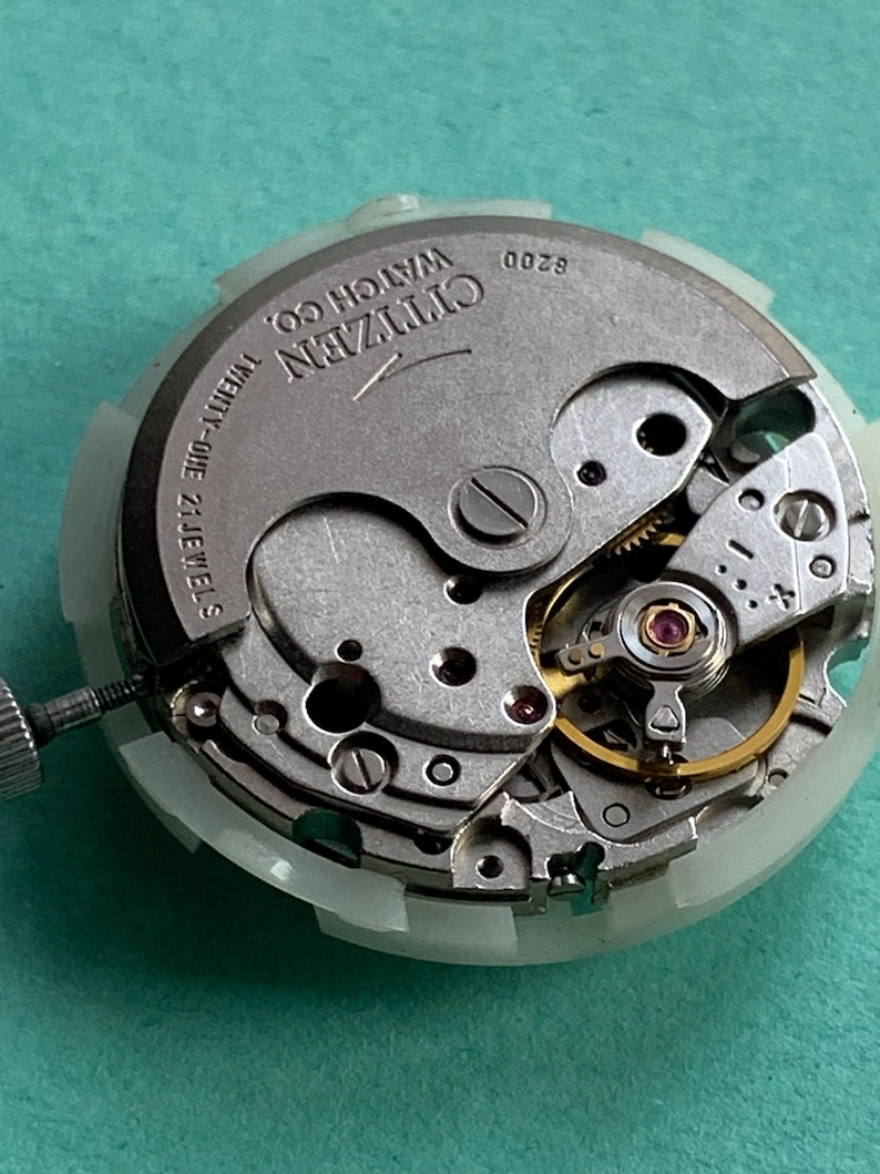 May include: Close-up of a Citizen watch movement with visible gears, jewels, and the text "Citizen Watch Co." The movement is silver-toned with a white plastic casing and a small screw. The watch movement is on a teal surface.