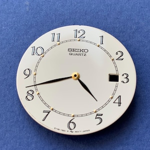 May include: A round, off-white Seiko Quartz watch dial featuring black Arabic numerals from 1 to 12, a black minute track with gold-tone hour markers, a date window at the 3 o'clock position, and black hands with a central gold-tone post.