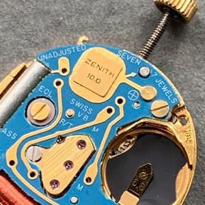 May include: Close-up of a Zenith watch movement with a blue circuit board and gold-colored components. The text "ZENITH 10.6" is visible, along with "SWISS V8" and "7 JEWELS". A gold crown is attached to the movement.