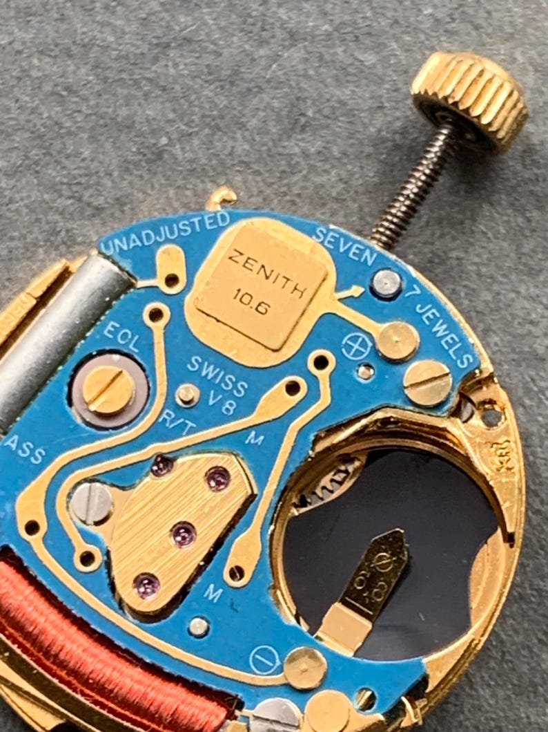 May include: Close-up of a Zenith watch movement with a blue circuit board and gold-colored components. The text "ZENITH 10.6" is visible, along with "SWISS V8" and "7 JEWELS". A gold crown is attached to the movement.
