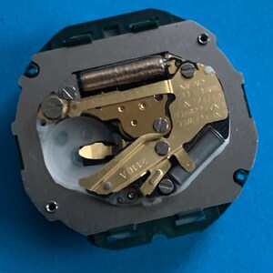 May include: A detailed view of a watch movement, featuring gold-toned gears, a silver plate, and a green plastic surround. The mechanism includes small screws and a coiled spring. The text "2110A" is visible.