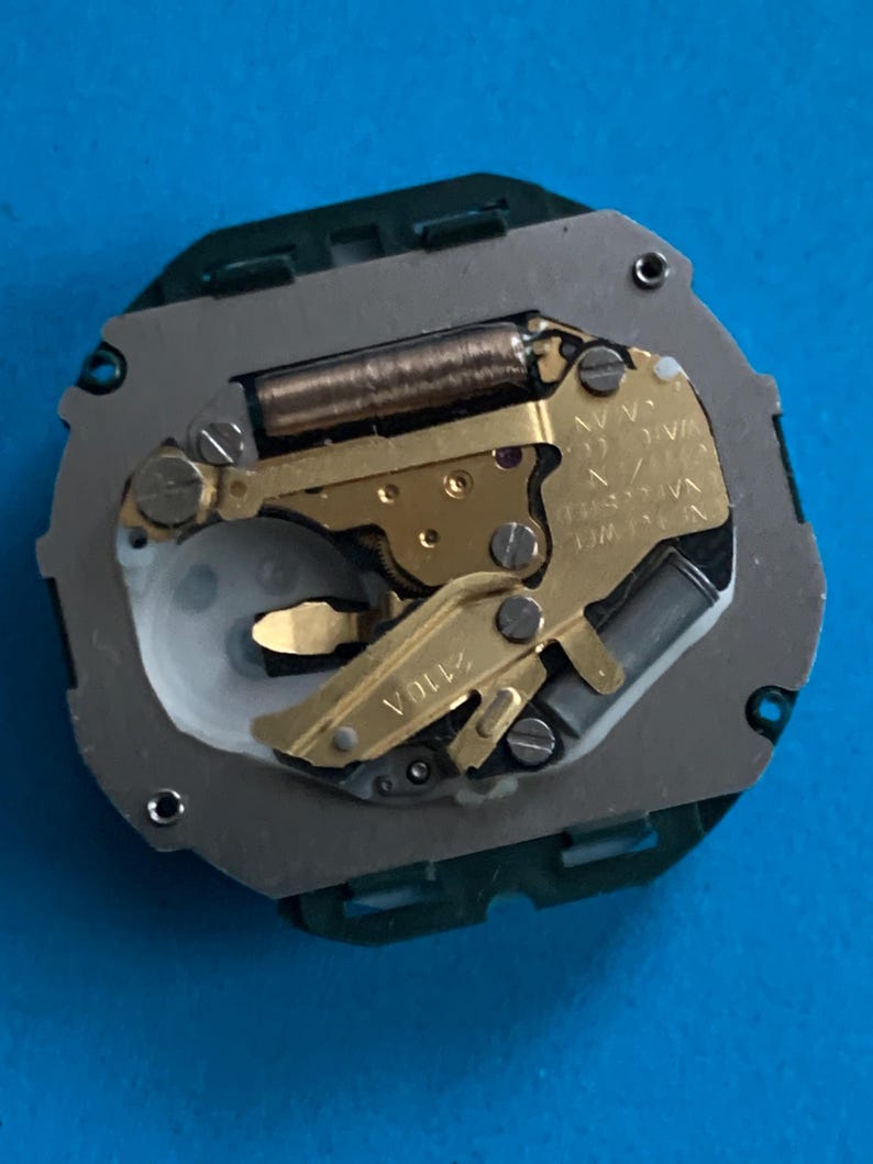 May include: A detailed view of a watch movement, featuring gold-toned gears, a silver plate, and a green plastic surround. The mechanism includes small screws and a coiled spring. The text "2110A" is visible.