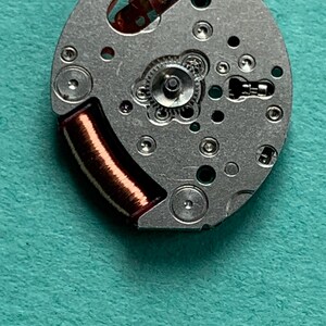 May include: Close-up of a watch movement with a silver-colored base. The movement features small gears, screws, and a copper-colored coil. The background is a teal-colored surface.