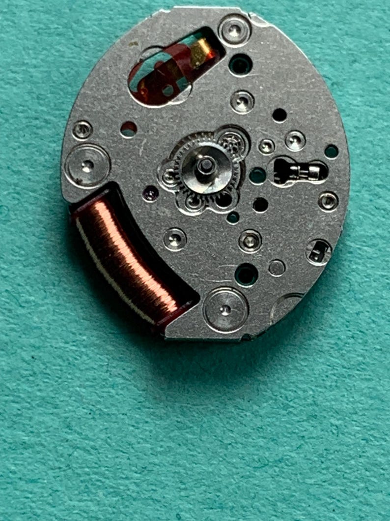 May include: Close-up of a watch movement with a silver-colored base. The movement features small gears, screws, and a copper-colored coil. The background is a teal-colored surface.