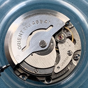 Op de afbeelding: Detailopname van een uurwerk met de tekst "ORIENT 21J 4694" en "JAPAN". Het mechanisme toont diverse tandwielen, schroeven en metalen componenten, die de ingewikkelde mechanische details laten zien.