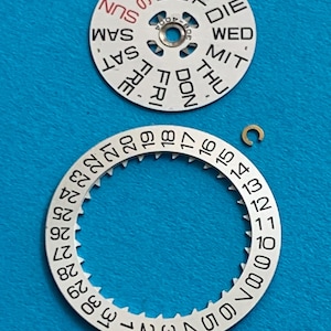 May include: Watch parts on a blue background. The image shows a white day-of-the-week dial with black text and a white date dial with black numbers. A small gold-colored clip is also present. The dials are components for a watch.
