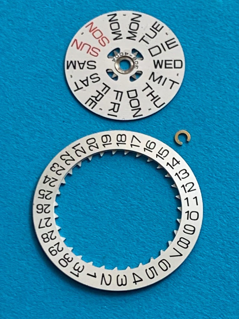 May include: Watch parts on a blue background. The image shows a white day-of-the-week dial with black text and a white date dial with black numbers. A small gold-colored clip is also present. The dials are components for a watch.