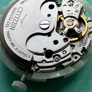 May include: Close-up of a Citizen watch movement, showcasing intricate mechanical components. The silver-toned mechanism features gears, jewels, and the text "Citizen Watch Co. Twenty-One 21 Jewels". A silver crown is visible.