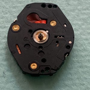 May include: A detailed view of a black watch movement, featuring intricate gears, gold-coloured studs, and a red element. The hexagonal component has several holes and fine details, revealing the internal workings of a watch.