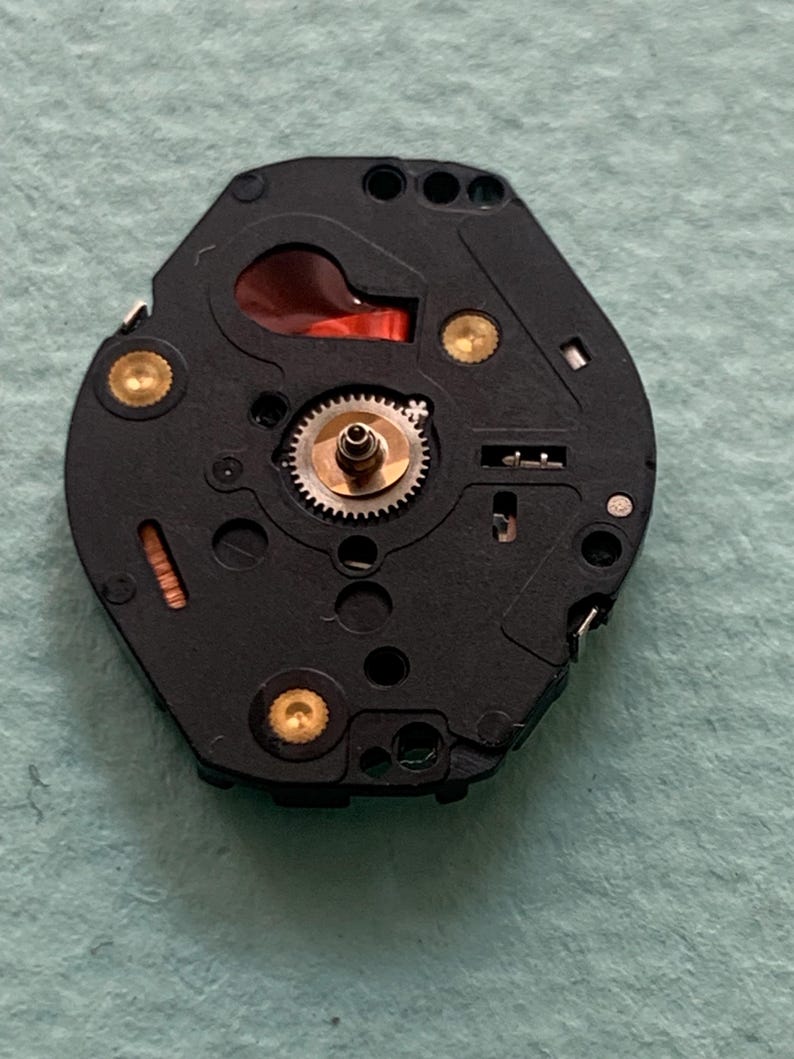 May include: A detailed view of a black watch movement, featuring intricate gears, gold-coloured studs, and a red element. The hexagonal component has several holes and fine details, revealing the internal workings of a watch.