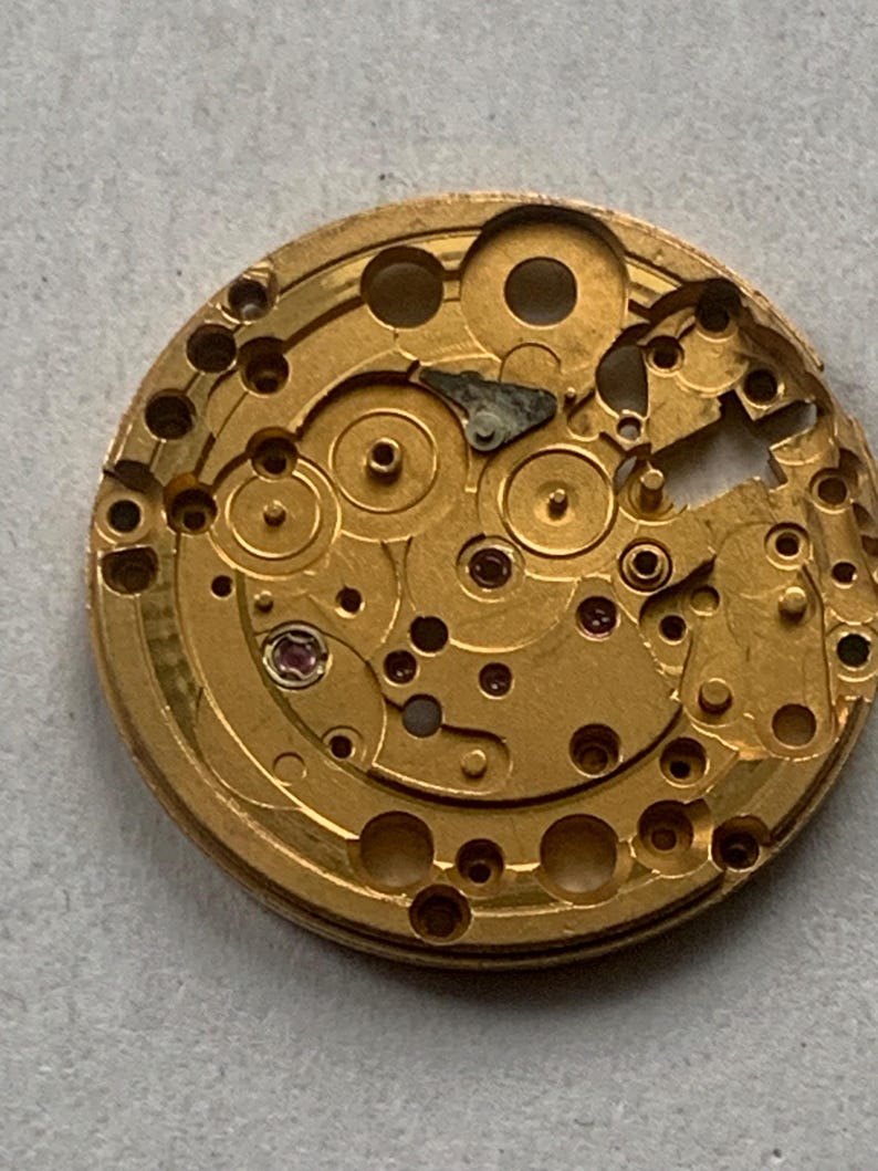 May include: Close-up of a gold-colored watch movement with intricate gears, wheels, and various components. The circular mechanism features numerous holes and a small, red jewel. The watch movement is a complex piece of machinery.