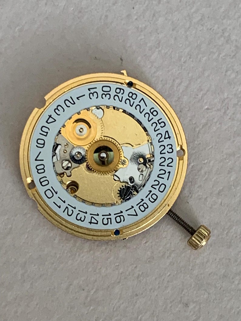 May include: A gold-toned watch movement with a light blue date dial. The dial features black numerals indicating the days of the month. A small screw and intricate internal gears are visible.