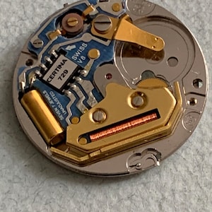 Może przedstawiać: Zbliżenie mechanizmu zegarka z misternymi detalami. Mechanizm zawiera niebieską płytkę drukowaną z napisem "Certina 729" i "Swiss V8". Widoczne są złote elementy i srebrna metalowa obudowa. Złożone urządzenie mechaniczne.