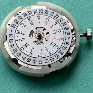 May include: A close-up of a watch movement with a white dial and silver-colored components. The dial displays the days of the week in English and Arabic, along with the date. A small crown is visible on the right side.